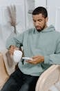 Young man in a hoodie using smartphone and holding coffee mug indoors, experiencing leisure time.